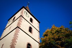 Nahaufnahme des Kirchturm der evangelischen Kirche Simmozheim vor blauem Himmel und neben einem Baum im Herbst