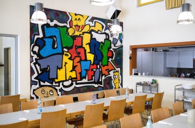Blick auf das bunte Graffiti im Raum des Jugendtreffs, im Vordergrund stehen Tischreihen
