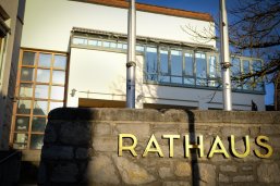 Schriftzug "Rathaus" auf einer Steinmauer, im Hintergrund ist der Sitzungssaal des Rathauses zu sehen