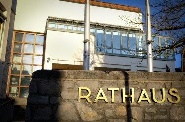 Schriftzug "Rathaus" auf einer Steinmauer, im Hintergrund ist der Sitzungssaal des Rathauses zu sehen