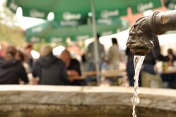 Brunnen im Vordergrund dahinter feiernde Menschen, die auf Bierbänken sitzen
